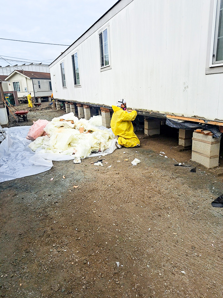 A person in a yellow hazmat suit works under a mobile home, removing insulation.