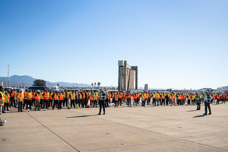 Large group of people wearing bright orange safety vests standing in formation outdoors.