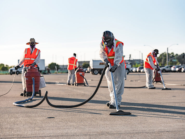 Four people in protective suits and masks use industrial vacuums to clean a paved surface.