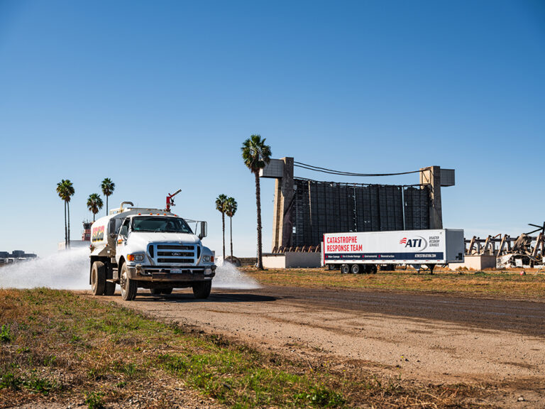 Water truck spraying water on a dirt road near a damaged building and a trailer labeled "Catastrophe Response Team."