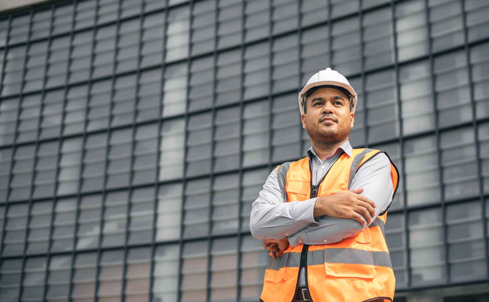 Construction worker in hard hat and safety vest with arms crossed, standing in front of a modern building.
