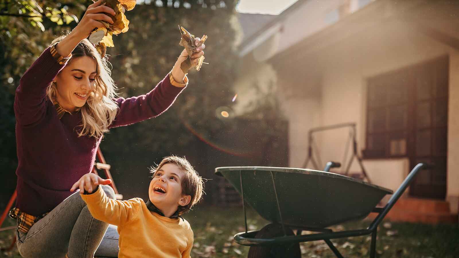 A woman and a child play with leaves in the backyard, with a wheelbarrow in the background.