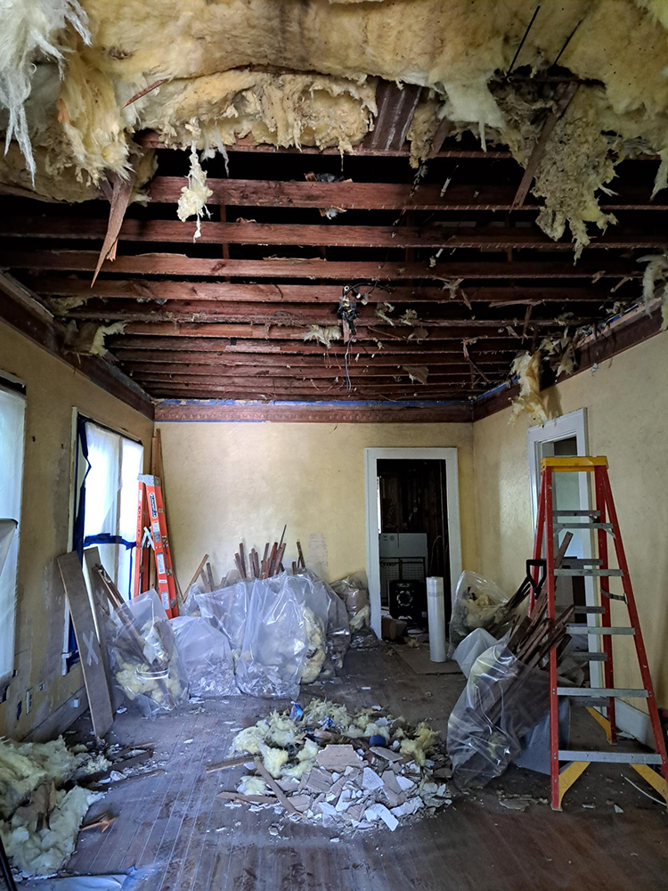 A room undergoing demolition, with a collapsed ceiling, debris on the floor, and construction materials scattered around.