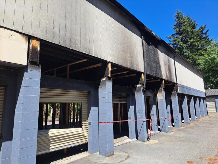 Fire-damaged row of garages with partially destroyed roll-up doors and caution tape.