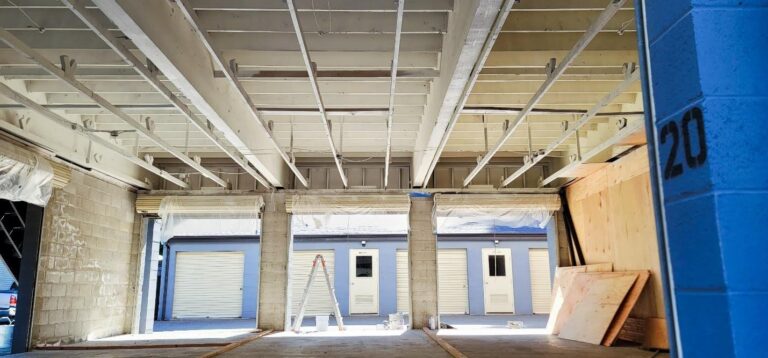 Interior view of a building under construction, showing an exposed ceiling with painted support beams, and several storage units with roll-up doors. A ladder and paint supplies are visible on the floor. A portion of a blue wall numbered "20" is in the foreground.