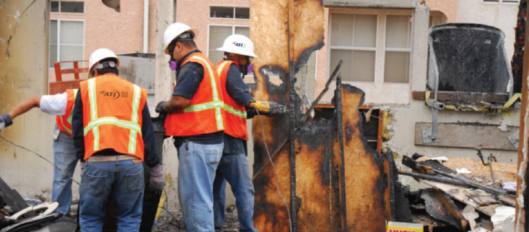 Three workers in hard hats and safety vests remove debris from a fire-damaged building.